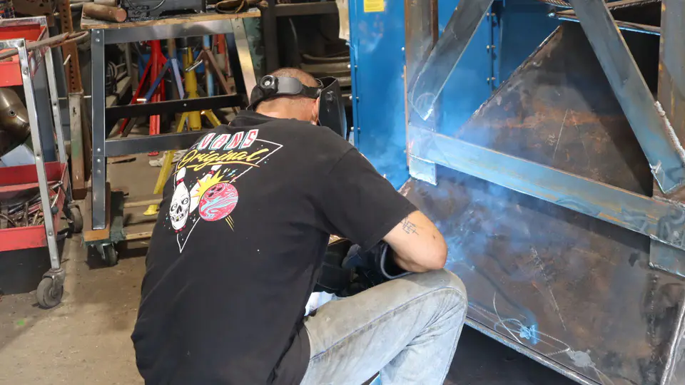A welder crouched at a big steel fabrication, arc light glowing blue